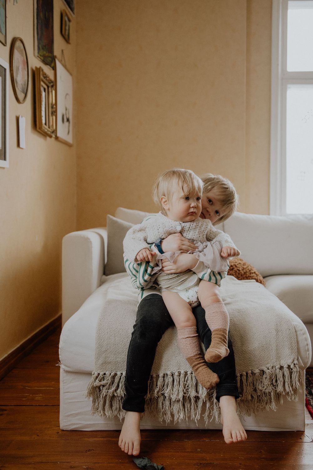 Two small children cuddle together on a white couch with fringe throw blanket in a sunlit room with hardwood floors.