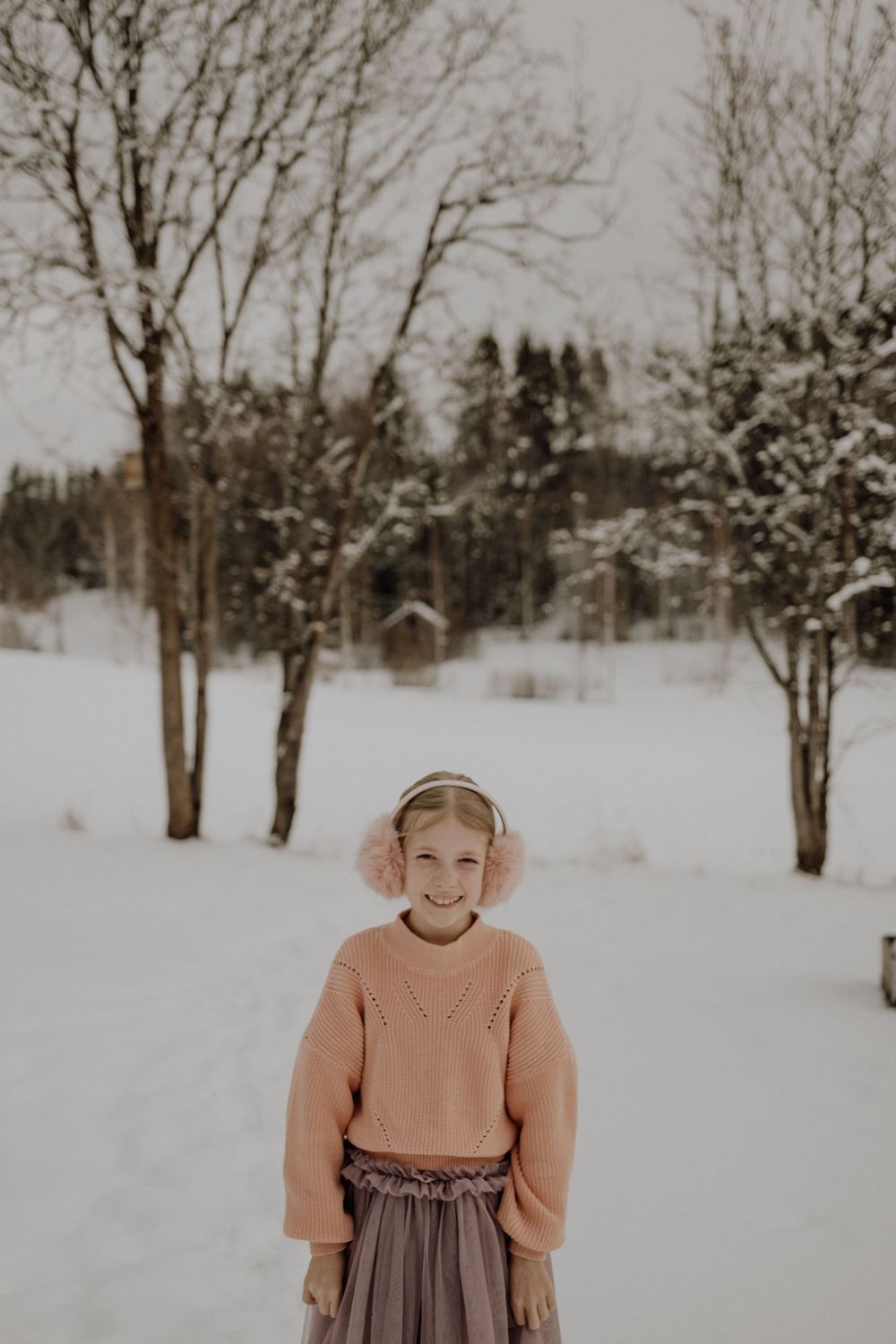 Person in pink earmuffs and peach sweater stands in snowy winter scene with bare trees in background.