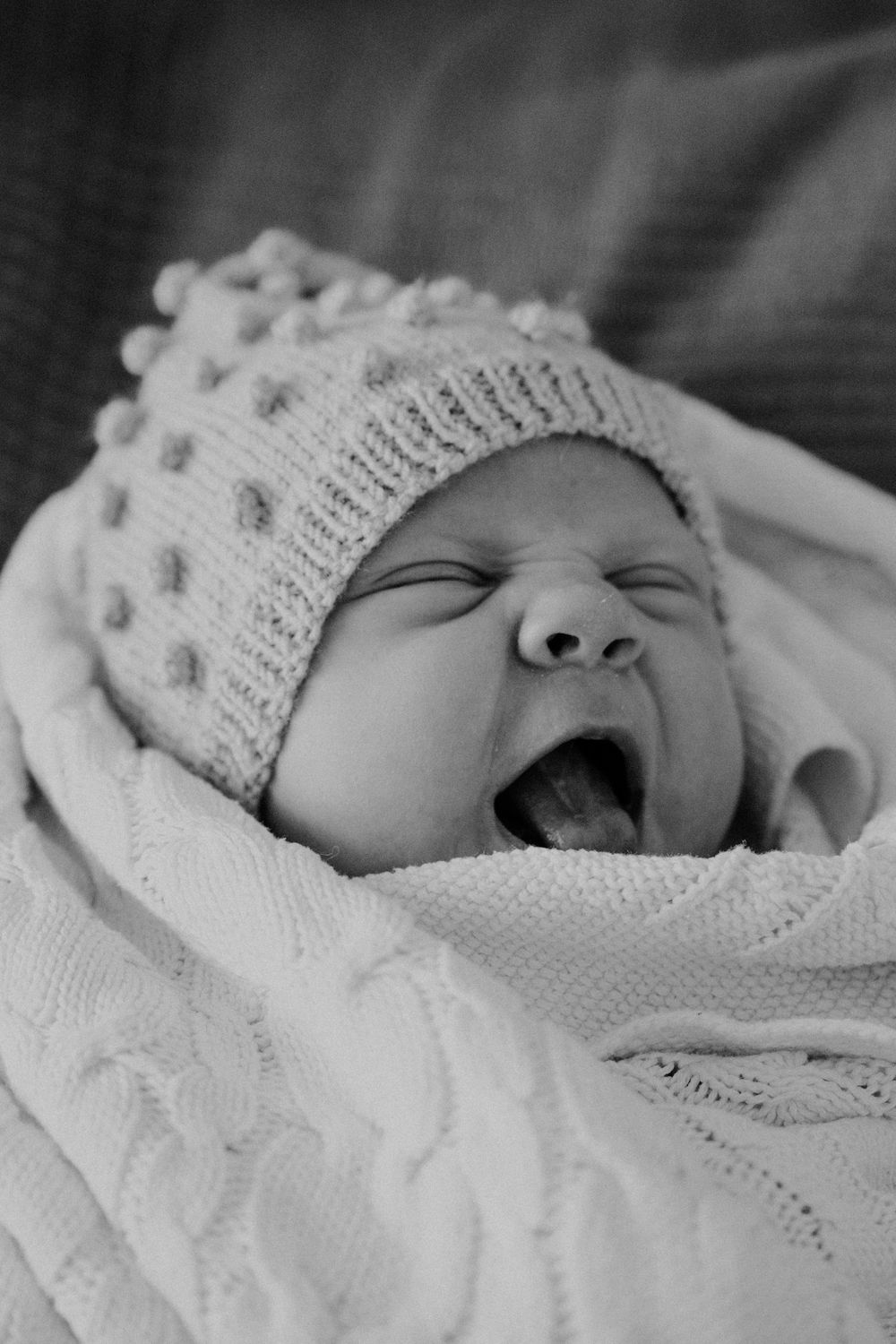 A tiny baby yawns while wearing a white knitted hat and wrapped in a soft blanket, captured in black and white.