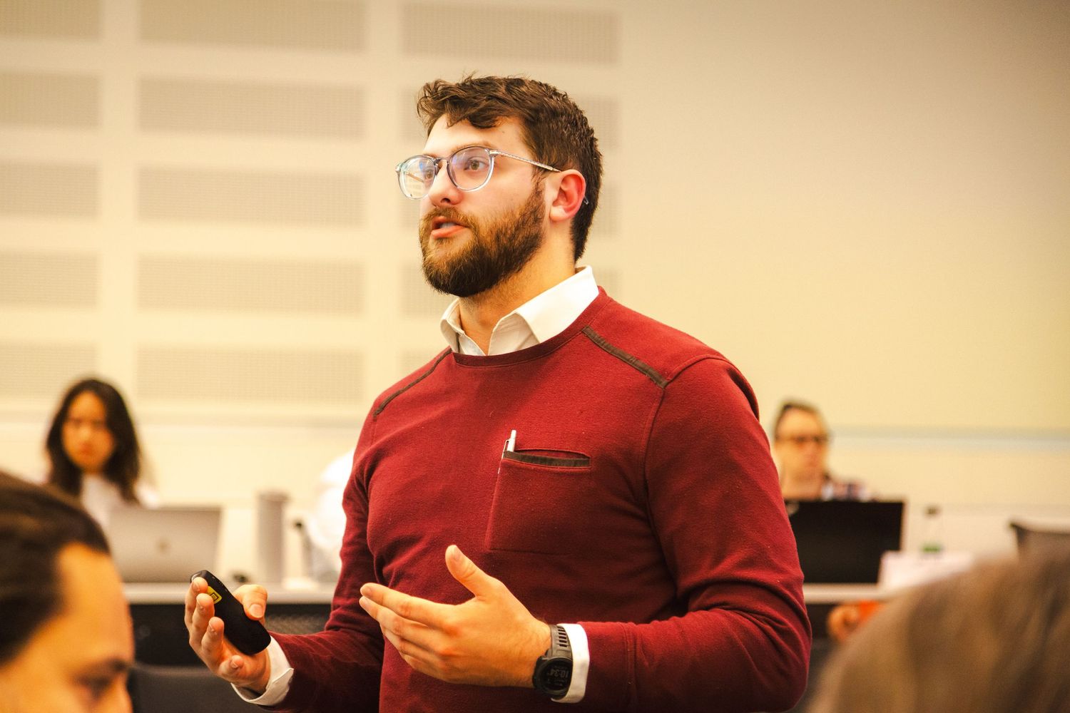 A speaker in a red sweater presents to an audience in a classroom setting.
