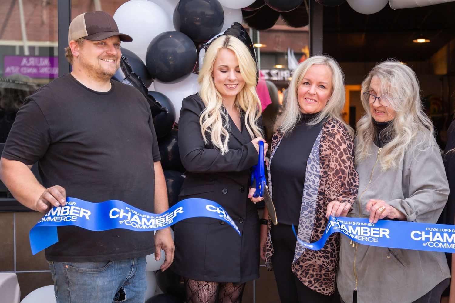 People holding blue ribbons prepare for a ceremonial ribbon cutting event with black and white balloons in the background.
