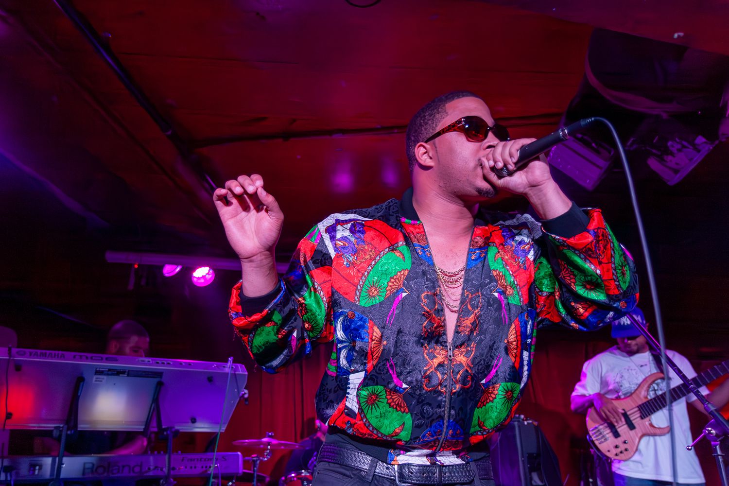 Performer in colorful tropical shirt sings on stage with microphone under dramatic purple lighting during live show.