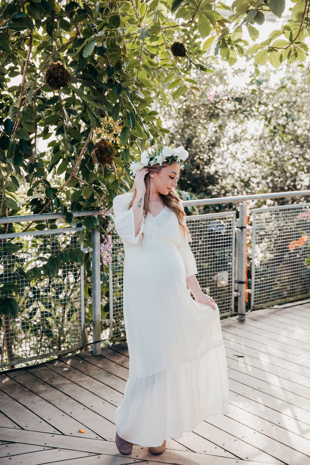Ethereal maternity photo of a woman in a flowing white dress and floral crown standing on a wooden deck.