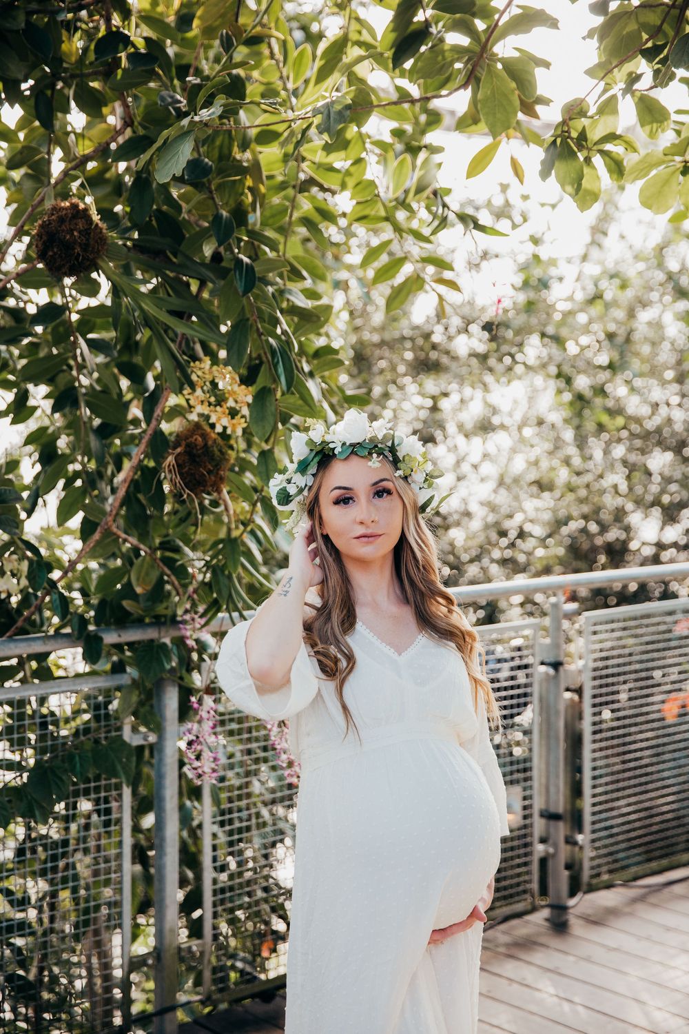Elegant maternity portrait with a woman in white dress and flower crown against lush greenery.