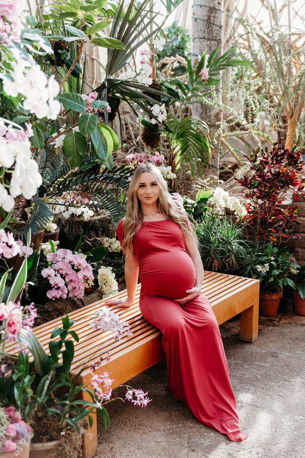 Maternity photo in a greenhouse setting with a woman in red dress surrounded by tropical plants.