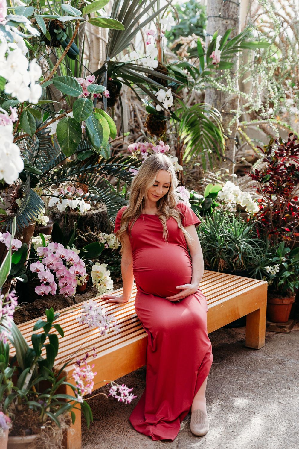 Expectant mother in red gown seated on wooden bench amidst pink flowers and tropical foliage.