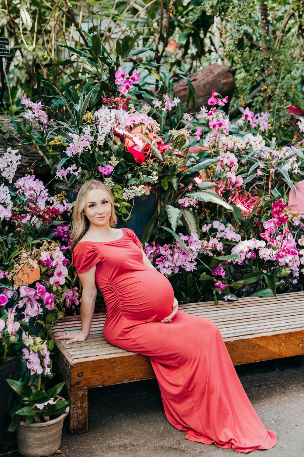 Artistic maternity photo with woman in coral dress reclining on bench surrounded by pink flowers.