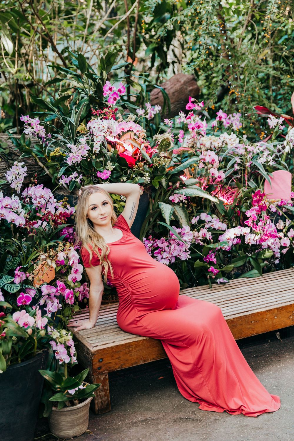 Romantic maternity photo with woman in red gown lounging among pink tropical flowers.
