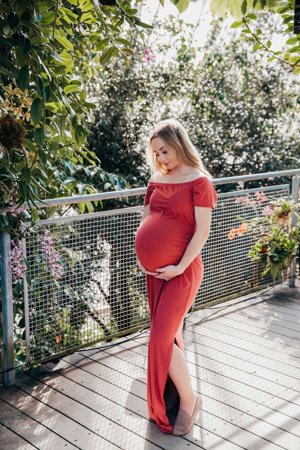 Side view maternity portrait of woman in red dress standing on outdoor deck with greenery.