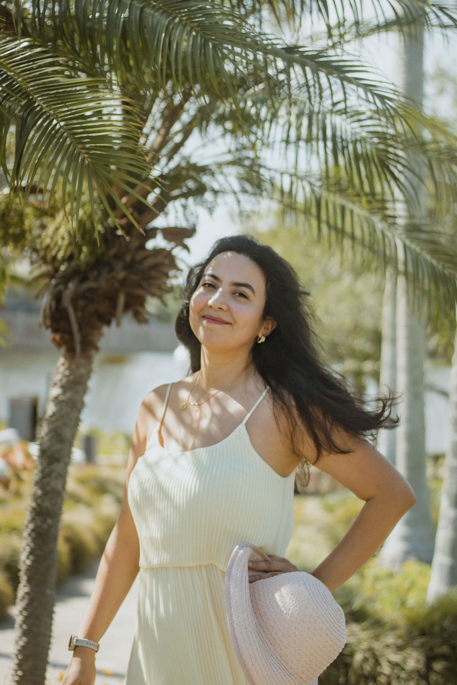 Young woman in white sundress holding a hat standing under palm trees on a sunny day.