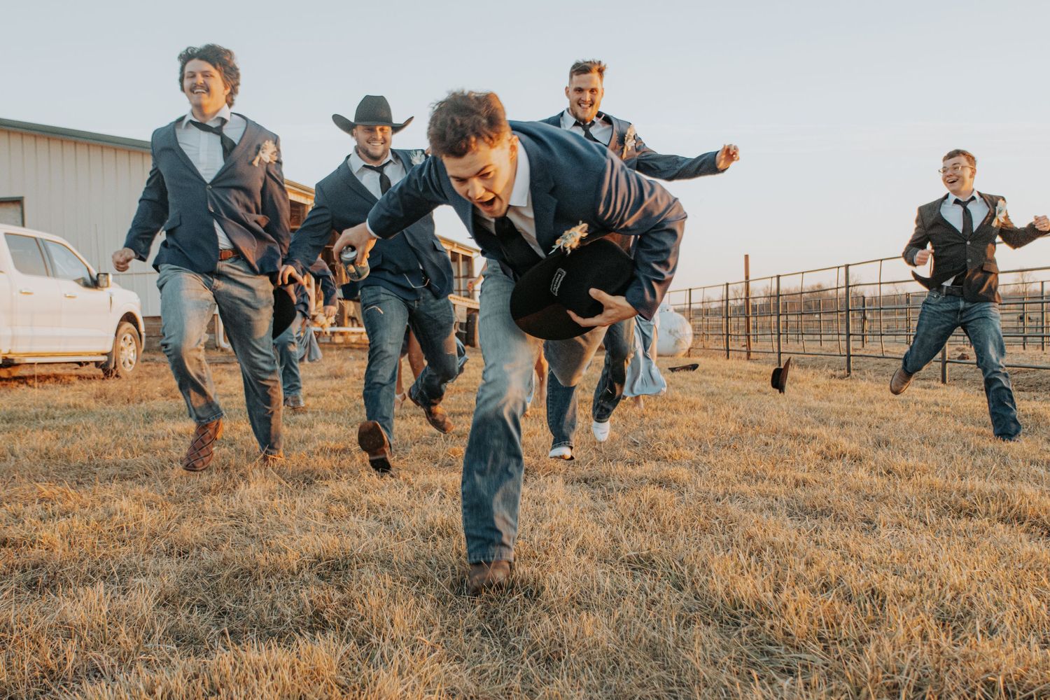 A bridal party in silver bridesmaid dresses and cowboy boots pose together on a rustic ranch at sunset.