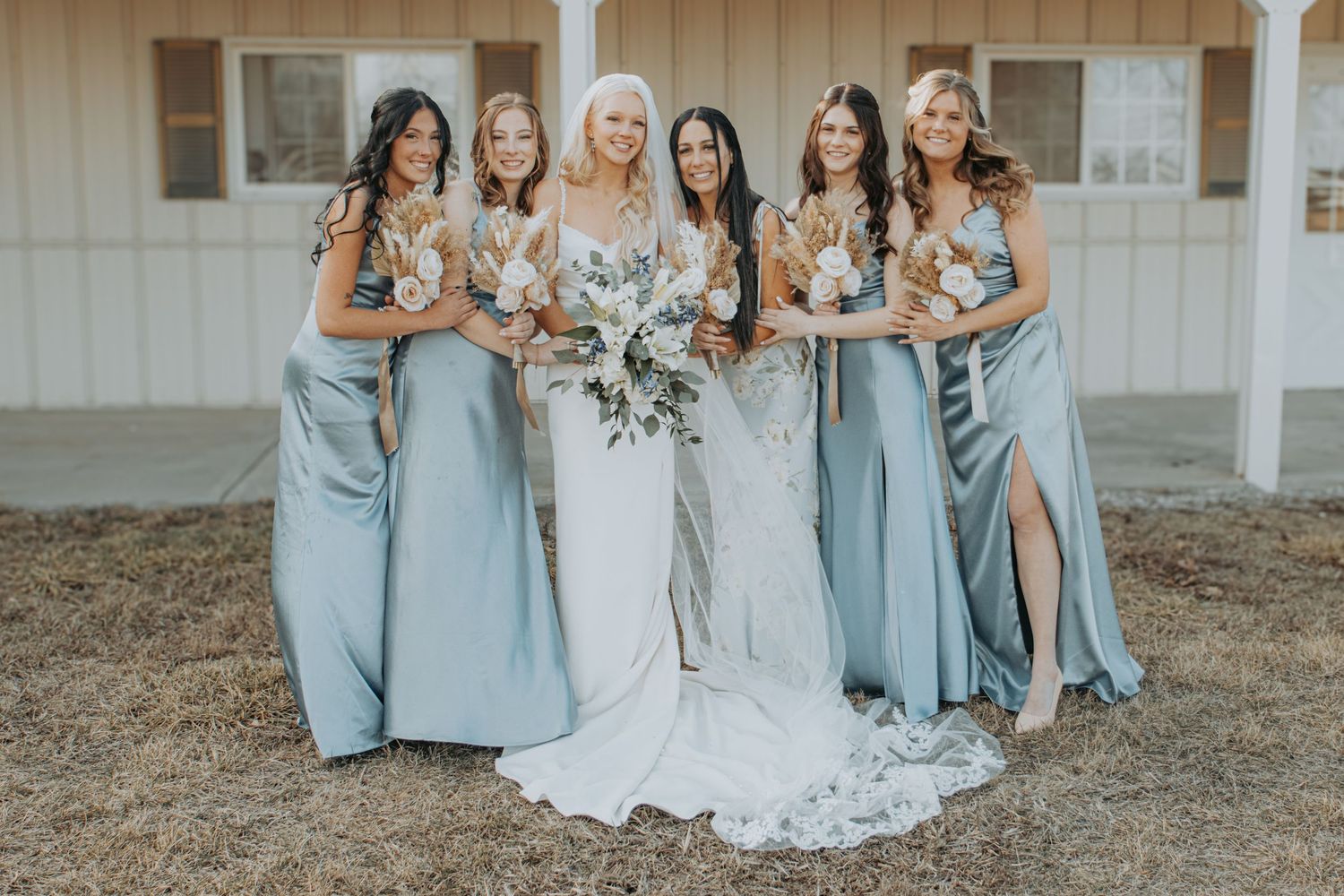A bridal party poses together outdoors wearing elegant dusty blue bridesmaid dresses and holding floral bouquets.