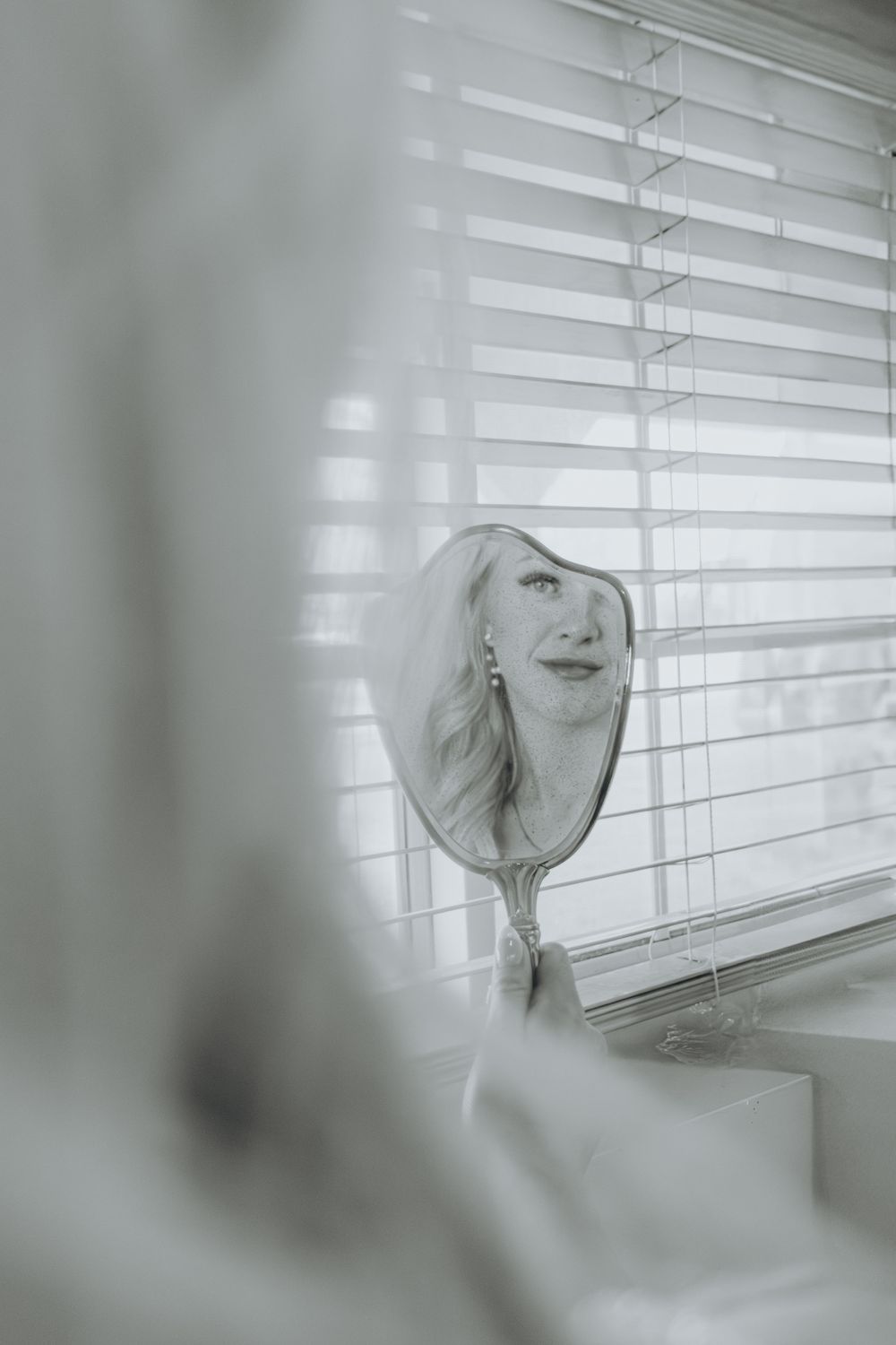 Artistic black and white mirror reflection showing long blonde hair and window blinds in soft focus.