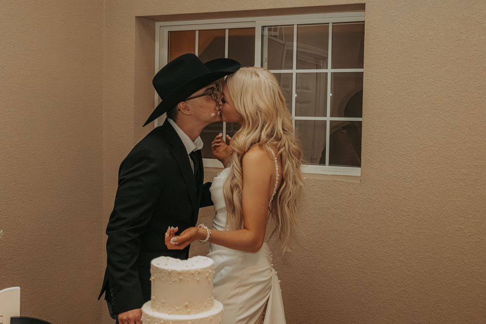 A romantic wedding cake cutting moment between a cowboy in black and bride in white at an indoor reception.