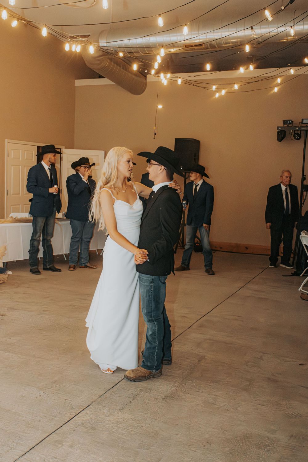 Couple shares first dance at wedding reception under string lights with guests watching in rustic venue.