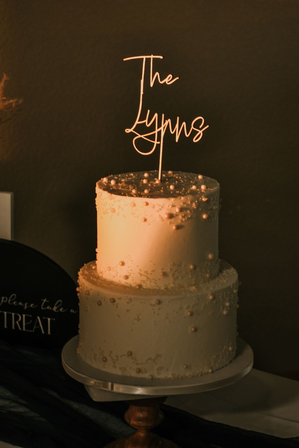 A two-tiered cream-colored wedding cake with gold accents and a glowing neon sign topper reading 'The Lynns'.