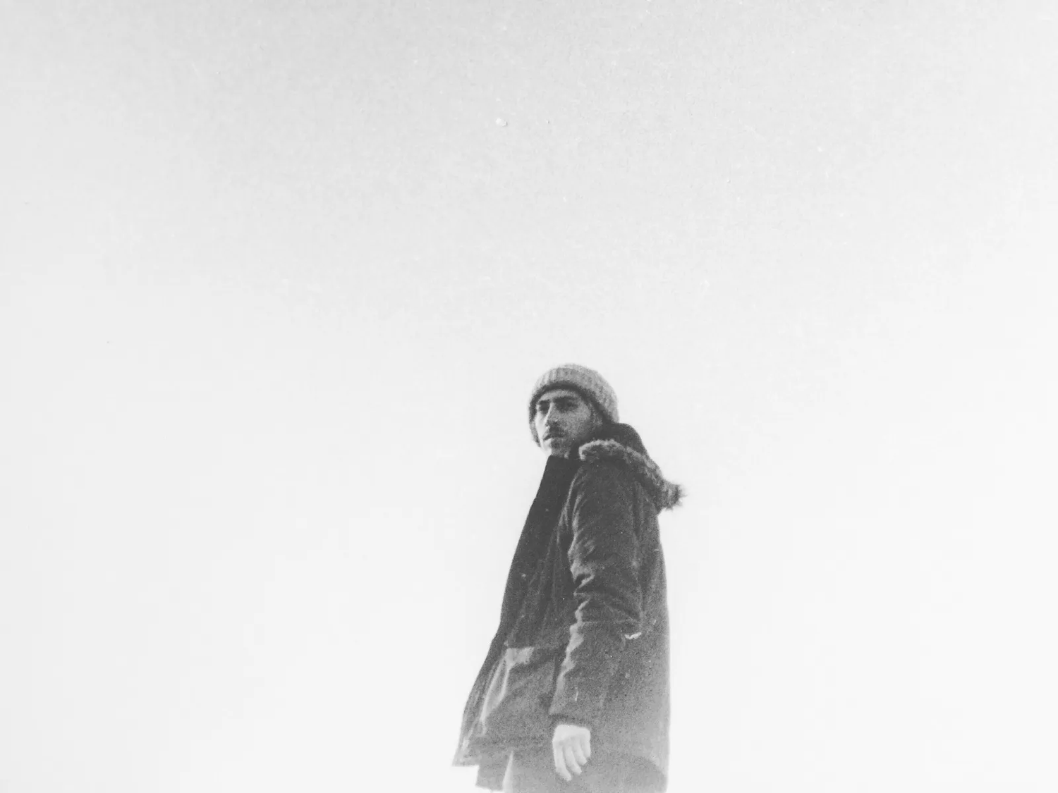 Femme en manteau sombre, debout seule contre un ciel surexposé, créant une silhouette détachée dans un décor presque blanc.