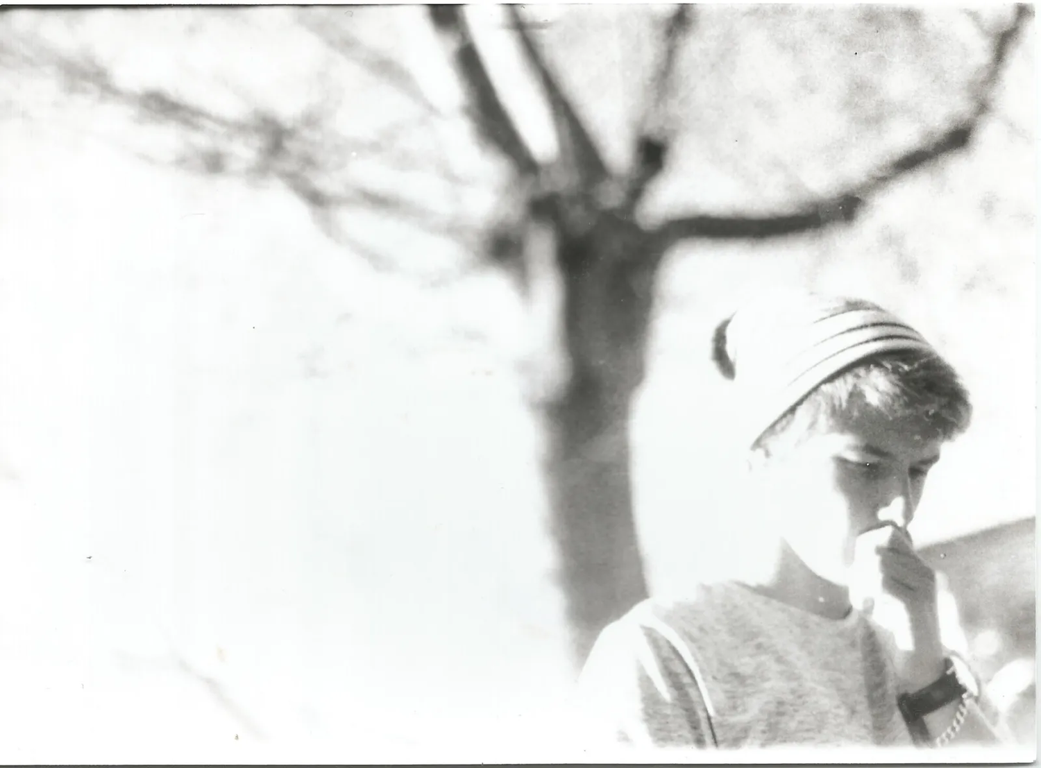 Portrait flou et surexposé d’un homme au bonnet clair, debout devant un arbre nu, dans une atmosphère fantomatique et poétique.