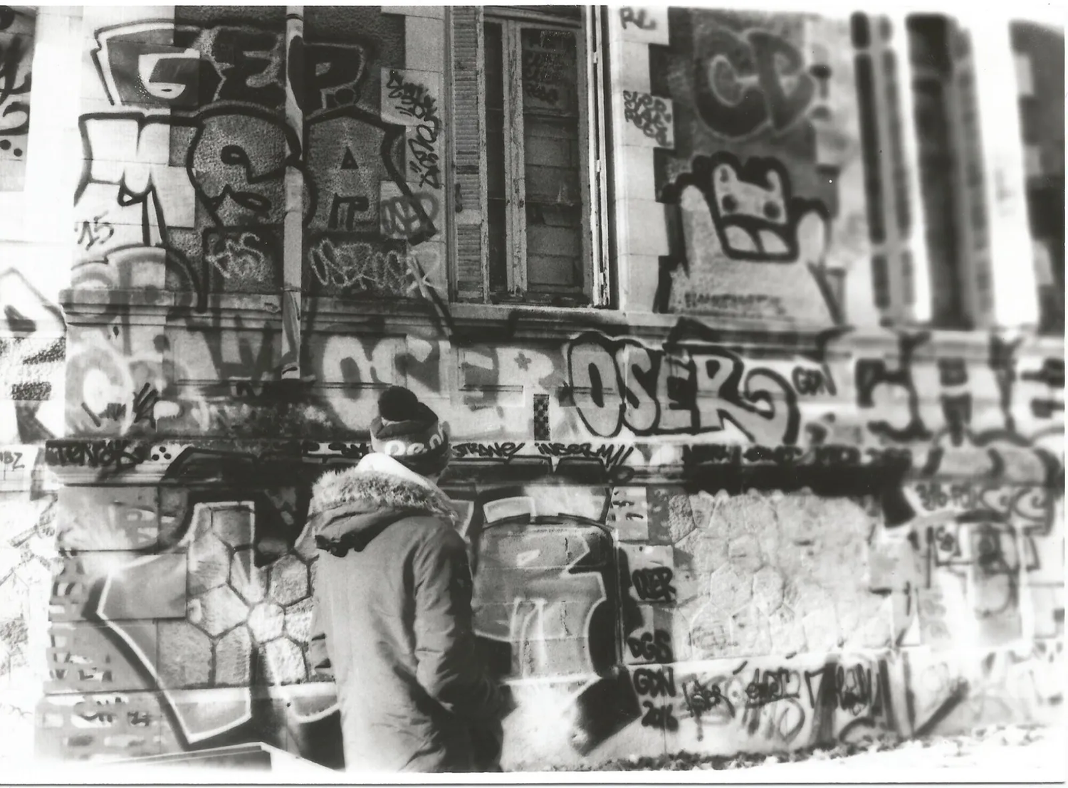 Mur urbain saturé de graffitis ; un individu se tient au centre, en partie camouflé dans la texture dense du décor.