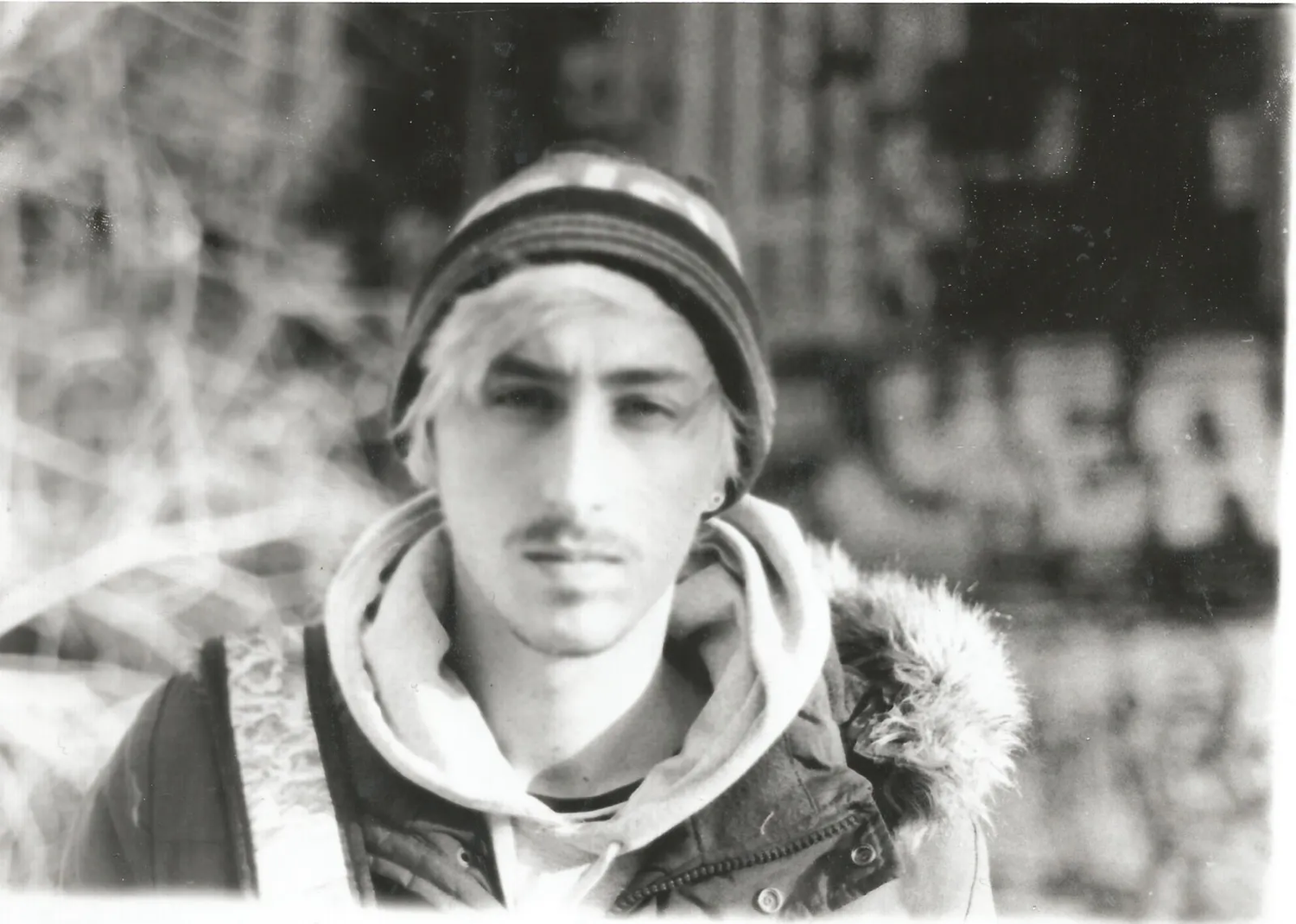 Portrait serré d’un jeune homme au regard franc, portant un bonnet et une parka, avec un mur tagué en arrière-plan.