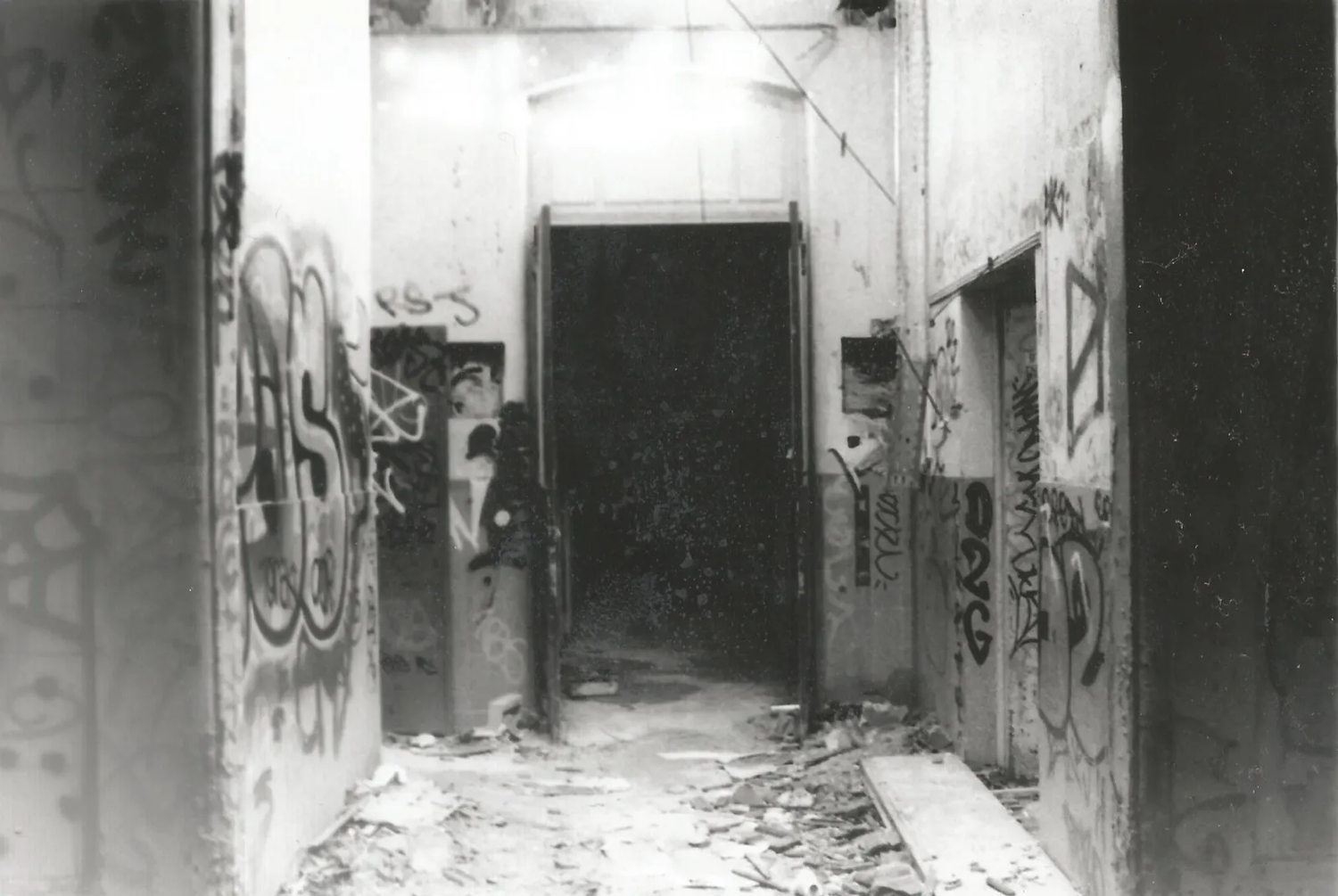 Couloir étroit aux murs couverts de graffitis, baigné d’une lumière surexposée en fond, créant une atmosphère urbaine abandonnée.