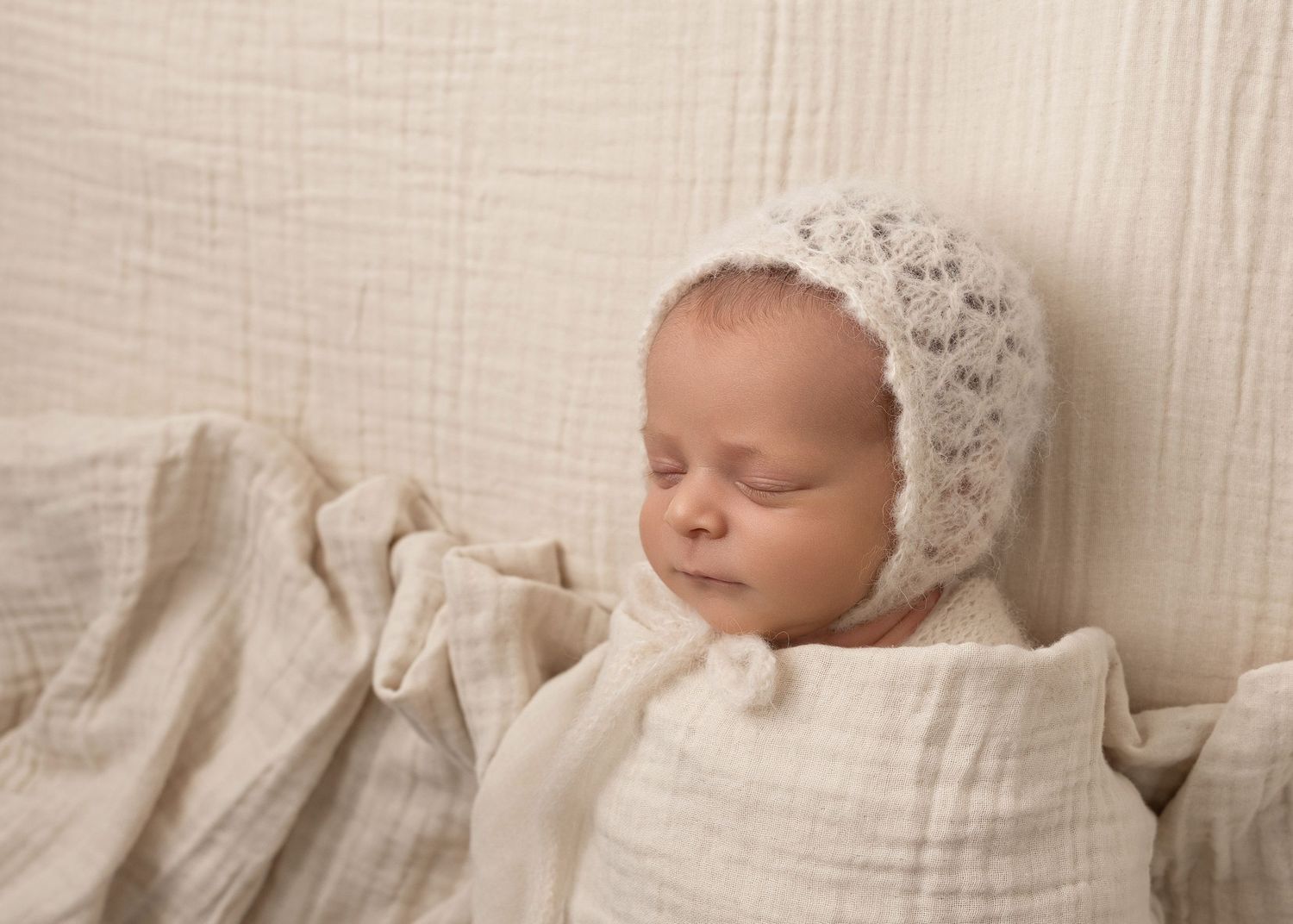 Sleeping newborn wearing delicate lace bonnet nestled in cream textured fabric.