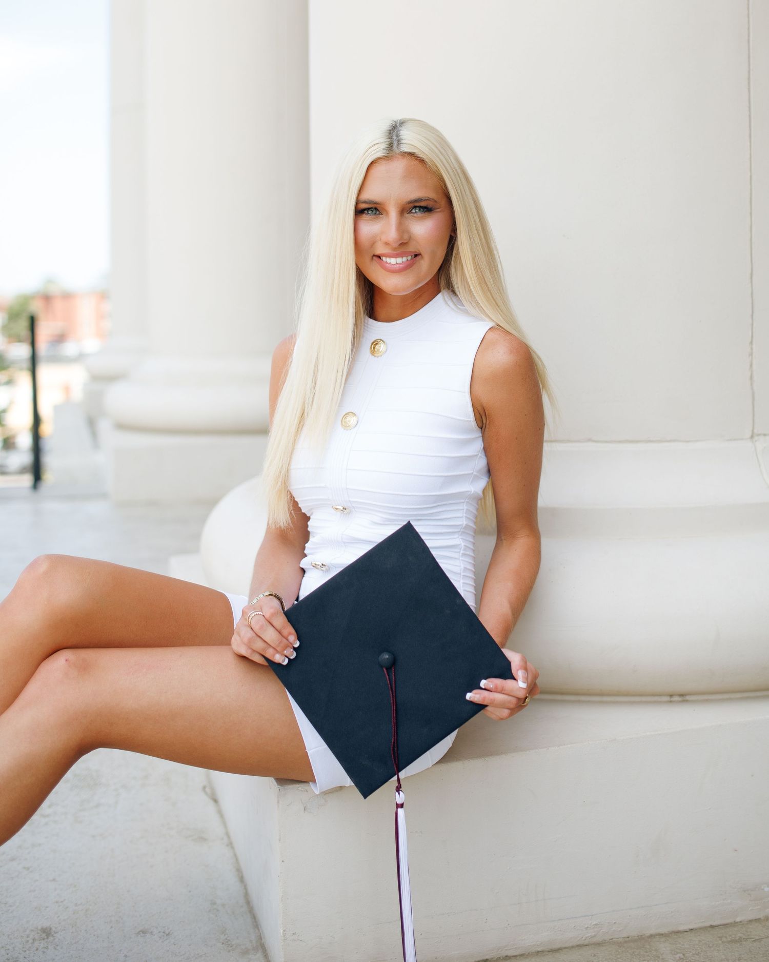 Graduate in white sleeveless dress poses with diploma against white column.