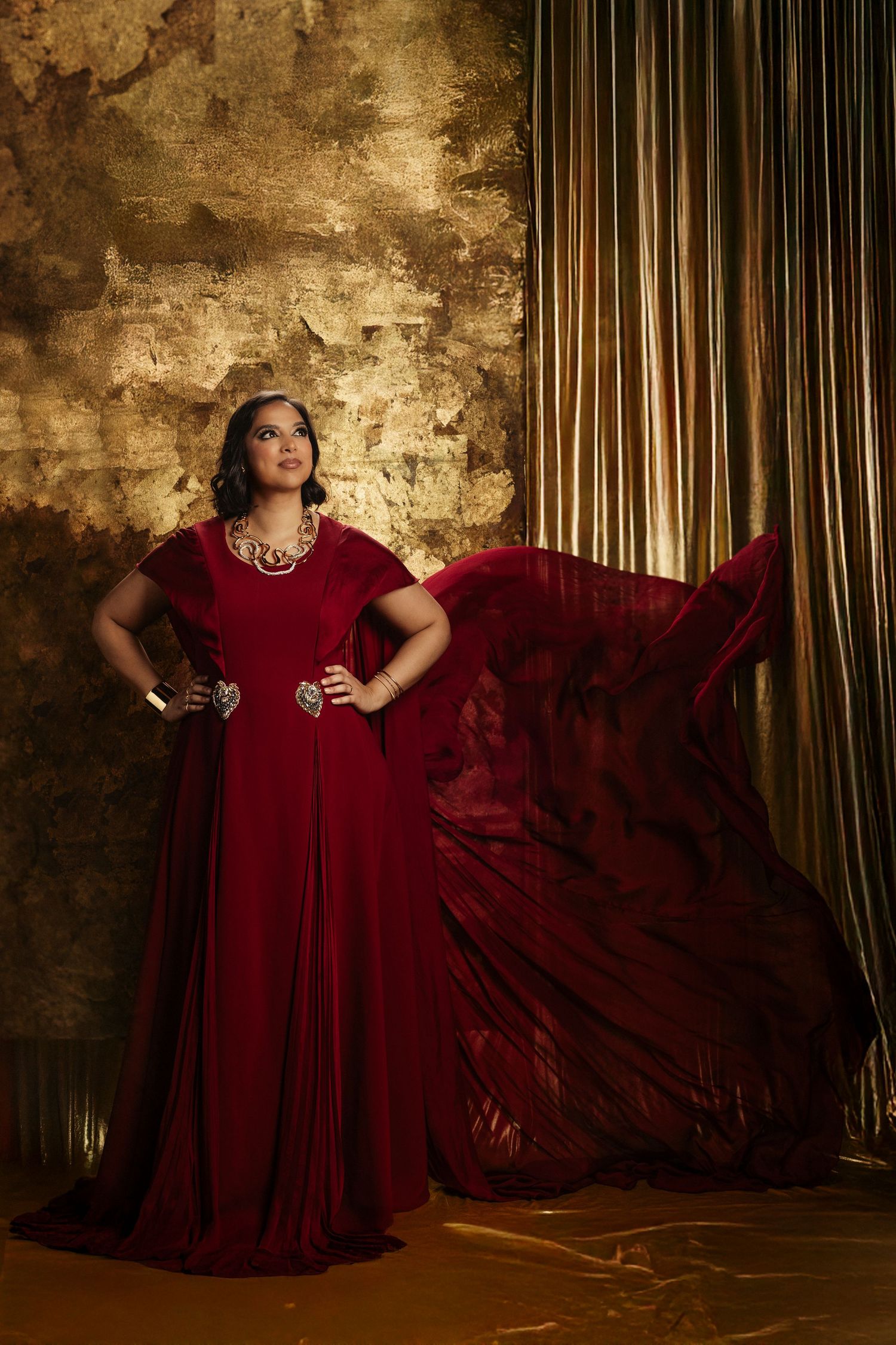 A dramatic portrait in a flowing red gown against golden textured walls and striped curtains.
