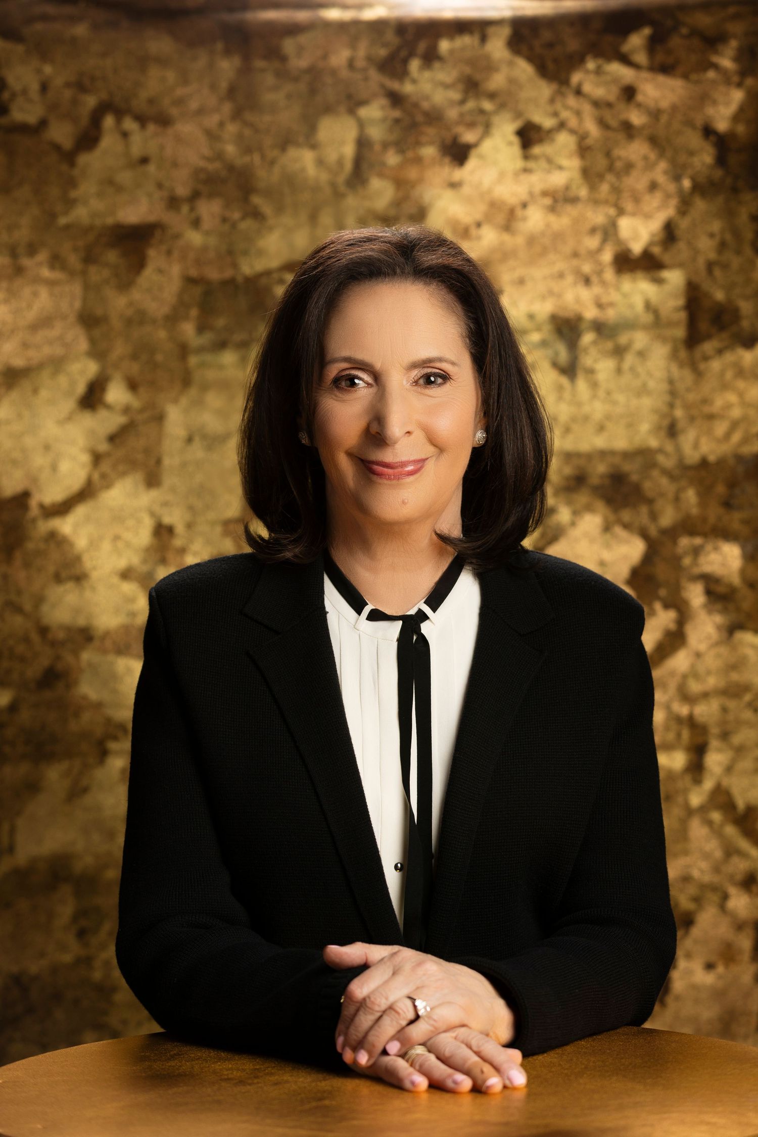 Professional studio portrait with black blazer and white blouse against textured gold backdrop.