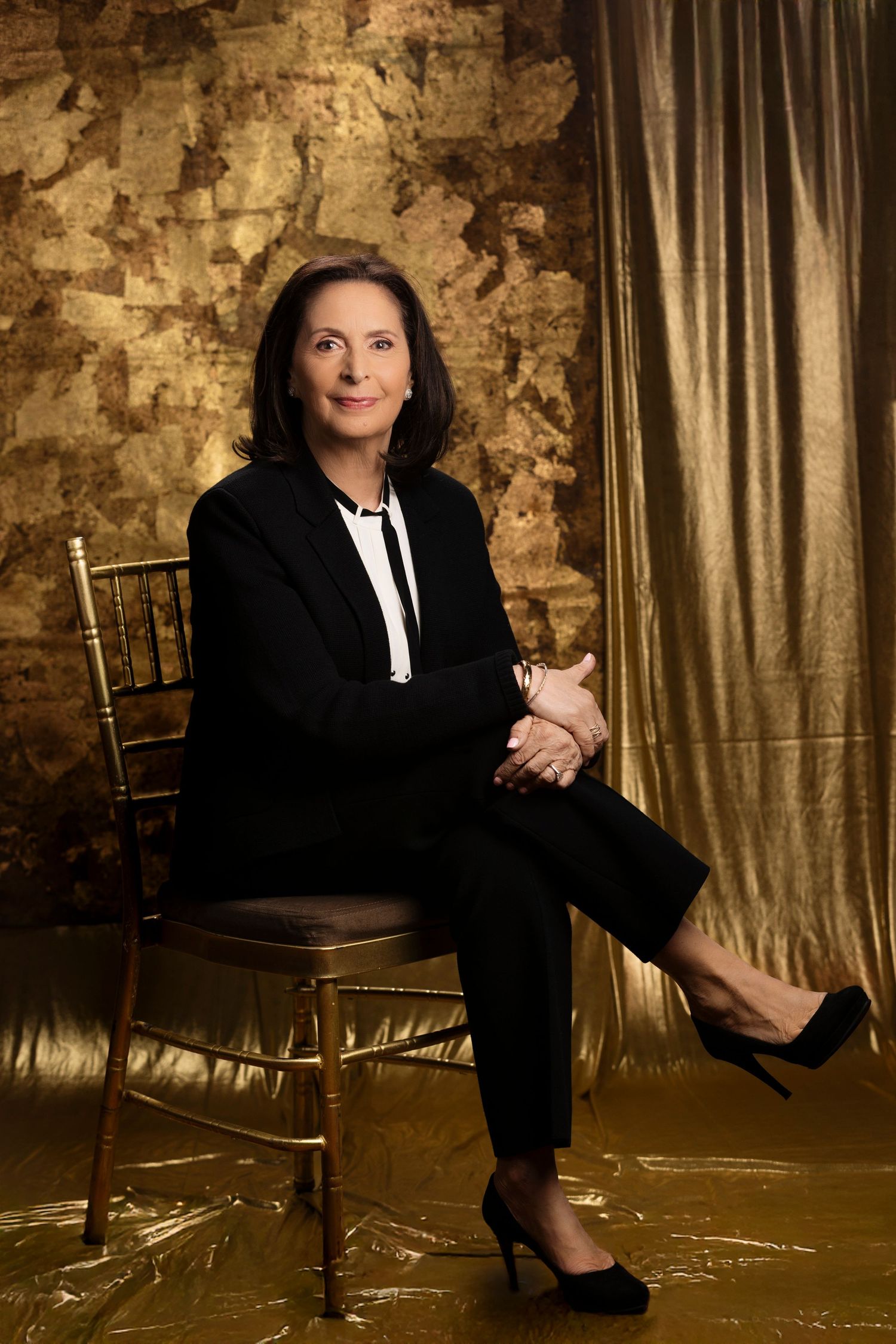 Professional portrait against textured gold wall showing a person in black business suit seated on gold chiavari chair.