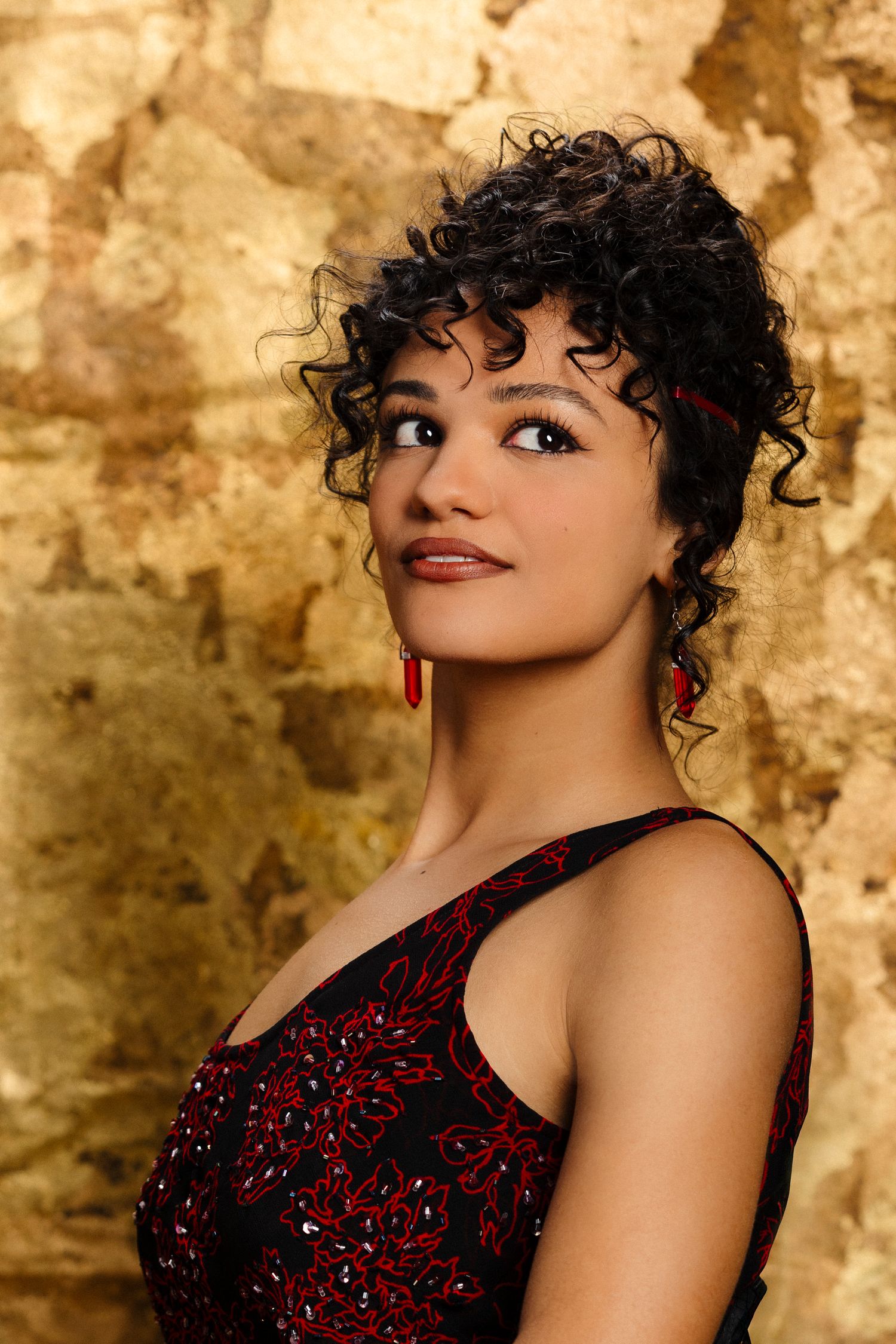Portrait of a person with curly dark hair wearing a red and black beaded dress against a textured golden background.