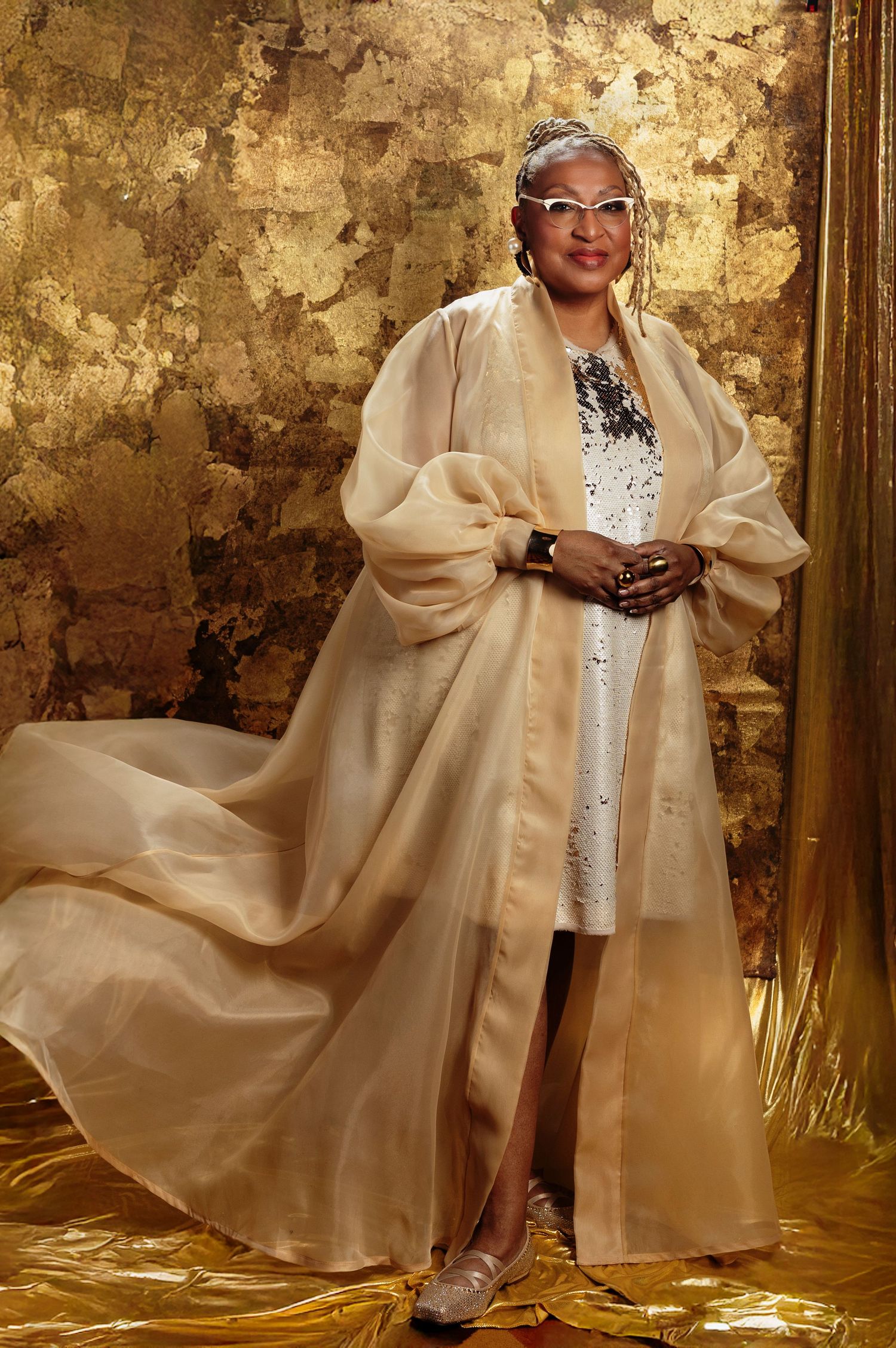 Elegant portrait in flowing cream coat and white dress against golden textured backdrop.
