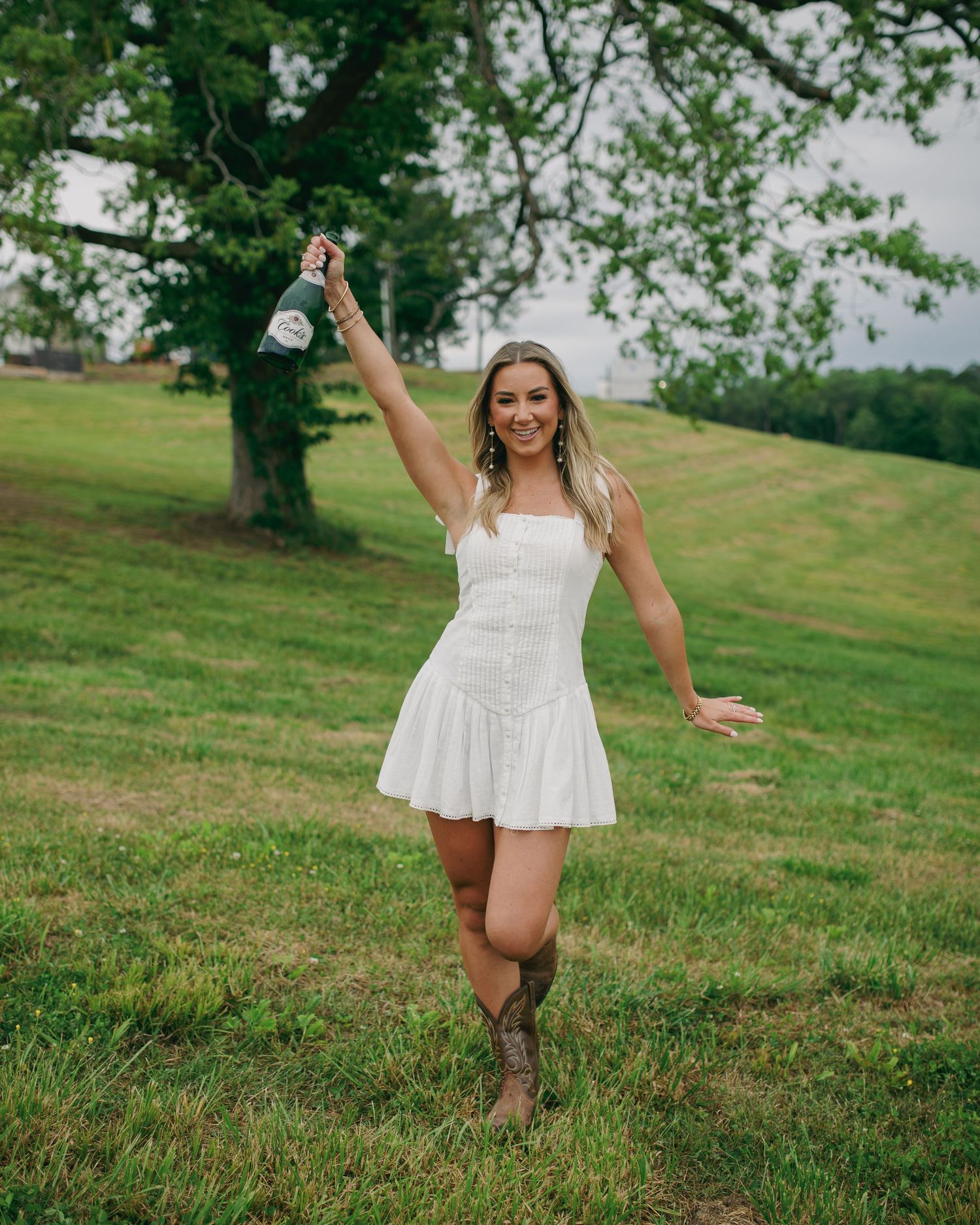 Woman in white dress and cowboy boots celebrates joyfully in a green field.