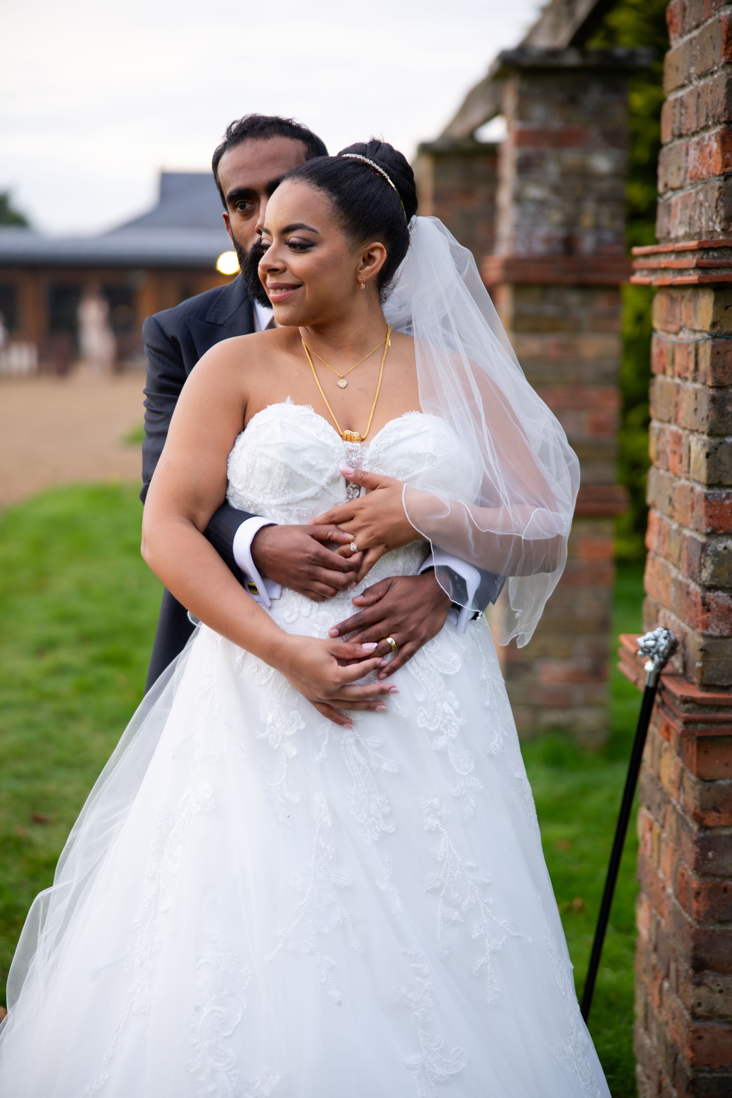 A couple embraces in a romantic outdoor wedding moment near a brick wall with the bride wearing a white strapless gown and veil.