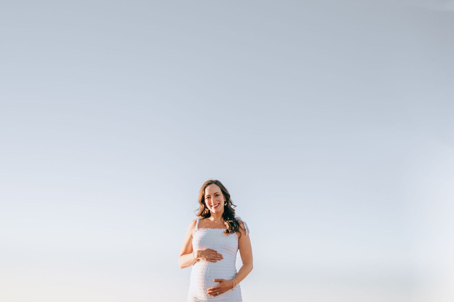 Maternity portrait of woman in white dress against minimal light background.