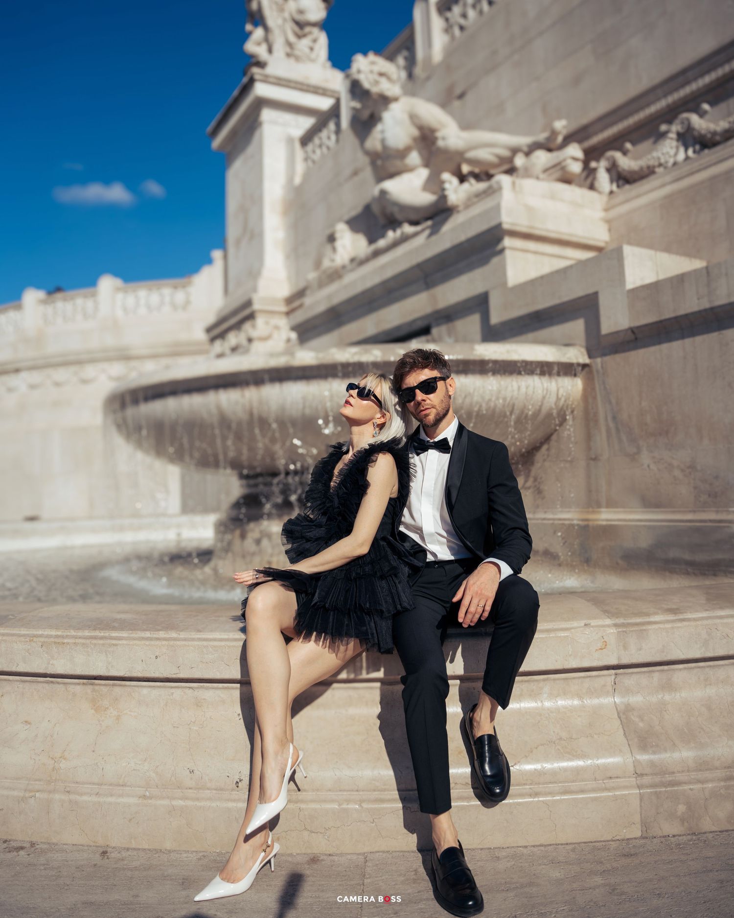 Stylish couple in formal attire pose together beside an ornate stone fountain on a sunny day.