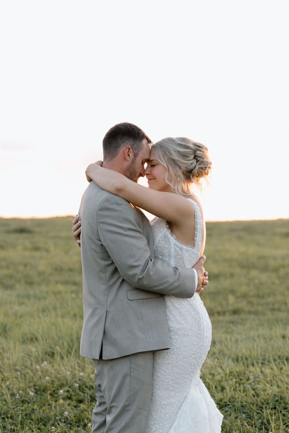 A romantic wedding couple embraces in a grassy field during golden hour.