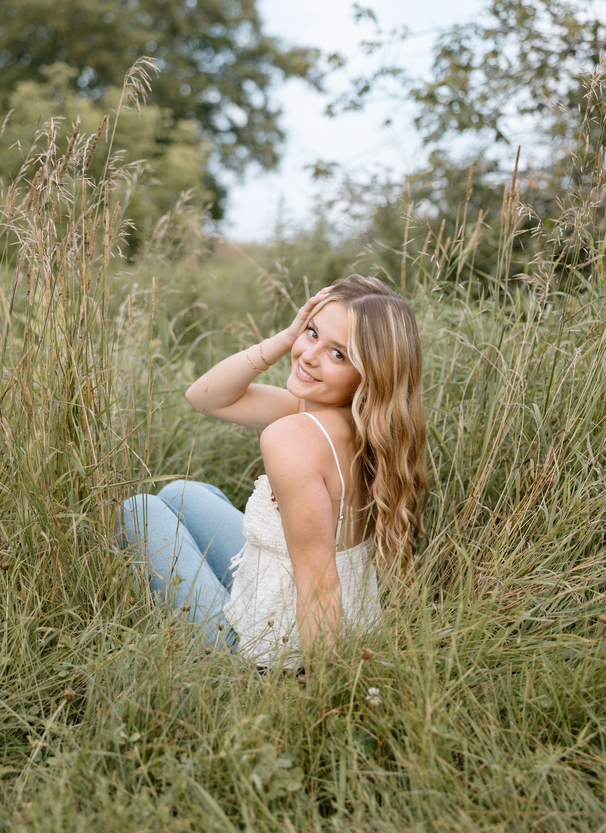 Senior in white top lying in tall summer grass during outdoor photo session.