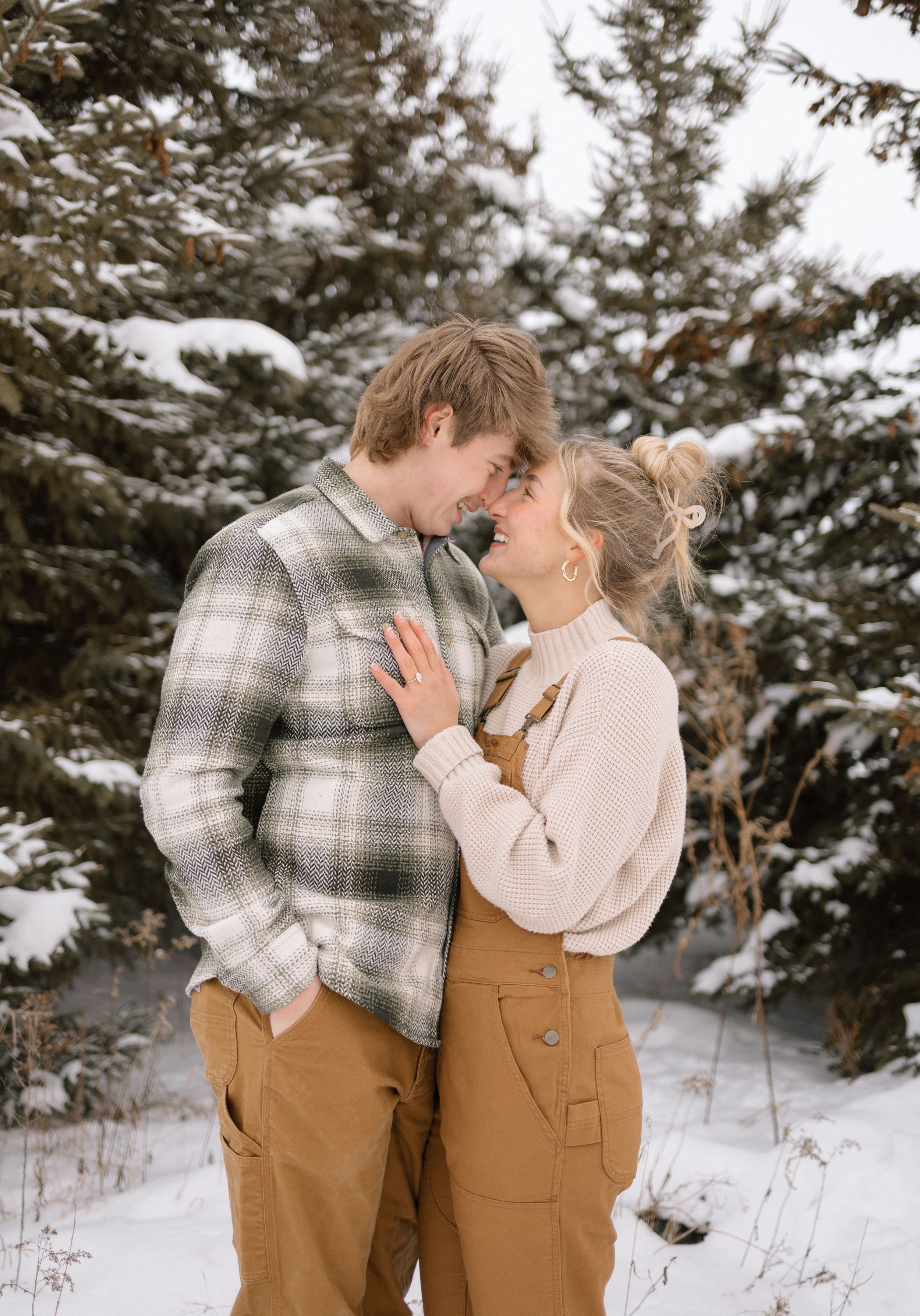 Romantic winter scene in Chippewa Falls, WI—couple in tan pants and sweaters hug in front of snowy evergreens by Ellie Rae Photography.
