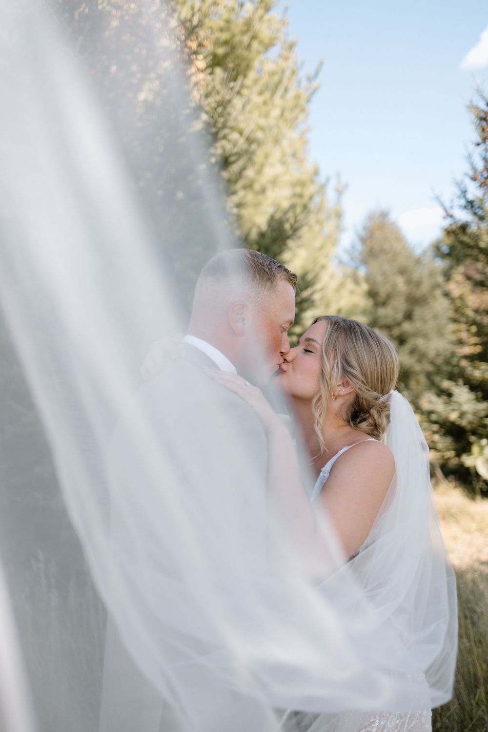 Ethereal bridal portrait with flowing white veil against evergreen trees.
