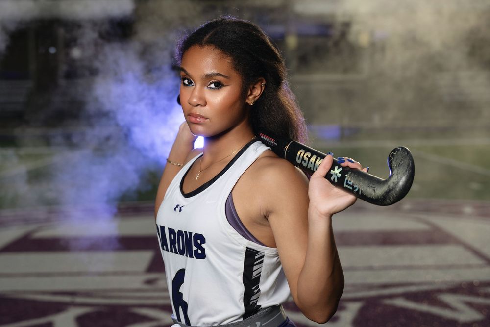 Field hockey athlete in white jersey poses with stick while blue smoke swirls in atmospheric sports portrait.