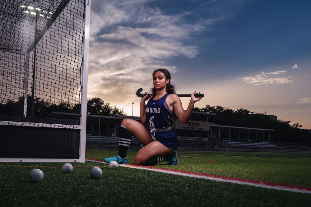 A soccer goalie in athletic gear sits on an artificial turf field at sunset with soccer balls scattered nearby.