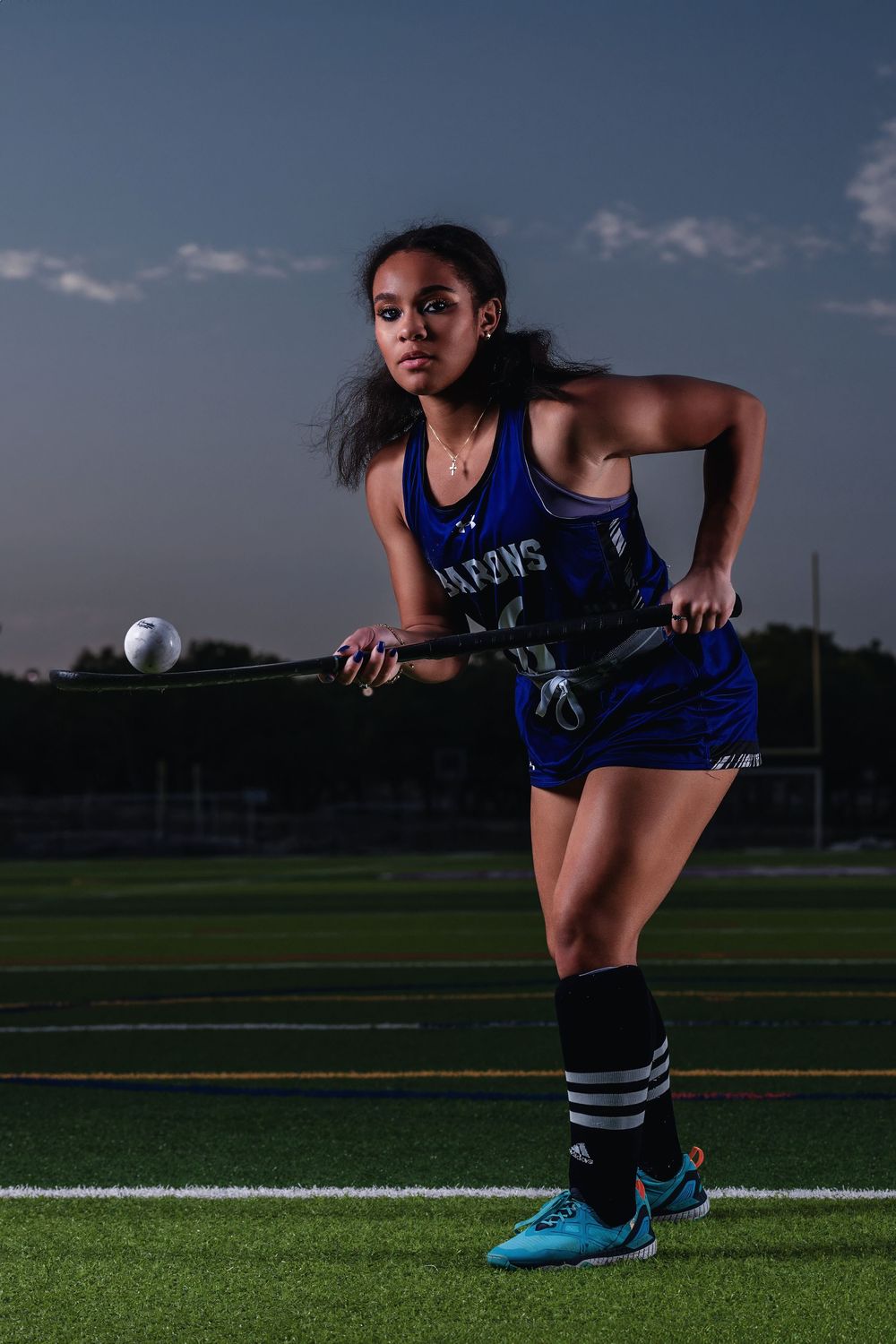 Athletic field hockey player in blue uniform crouches with stick ready for action on turf field at dusk.