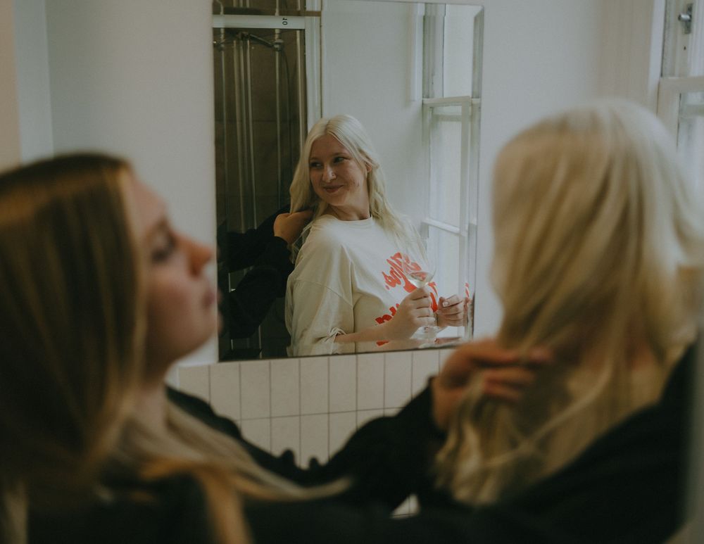 Three women getting ready in bathroom mirror wearing casual outfits with long blonde hair.