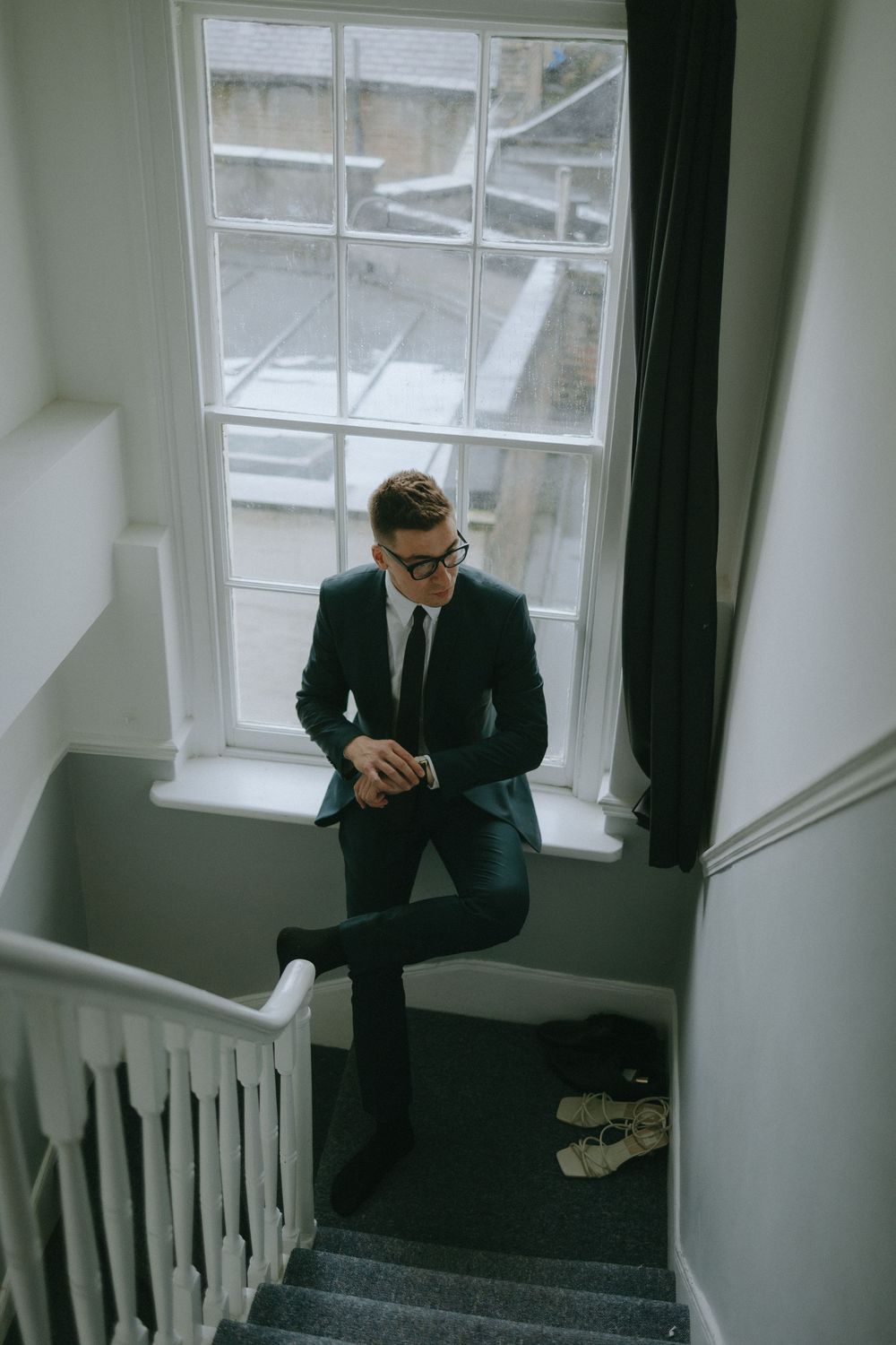 Person in dark suit sits on windowsill of elegant staircase landing with white walls.