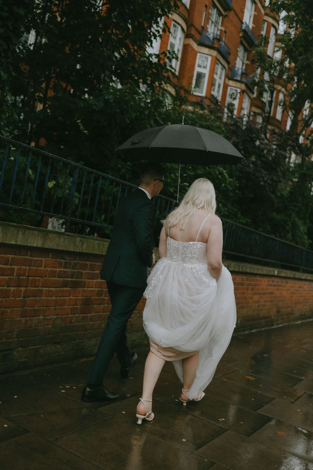 Couple walking in rain under black umbrella with bride holding up white dress.