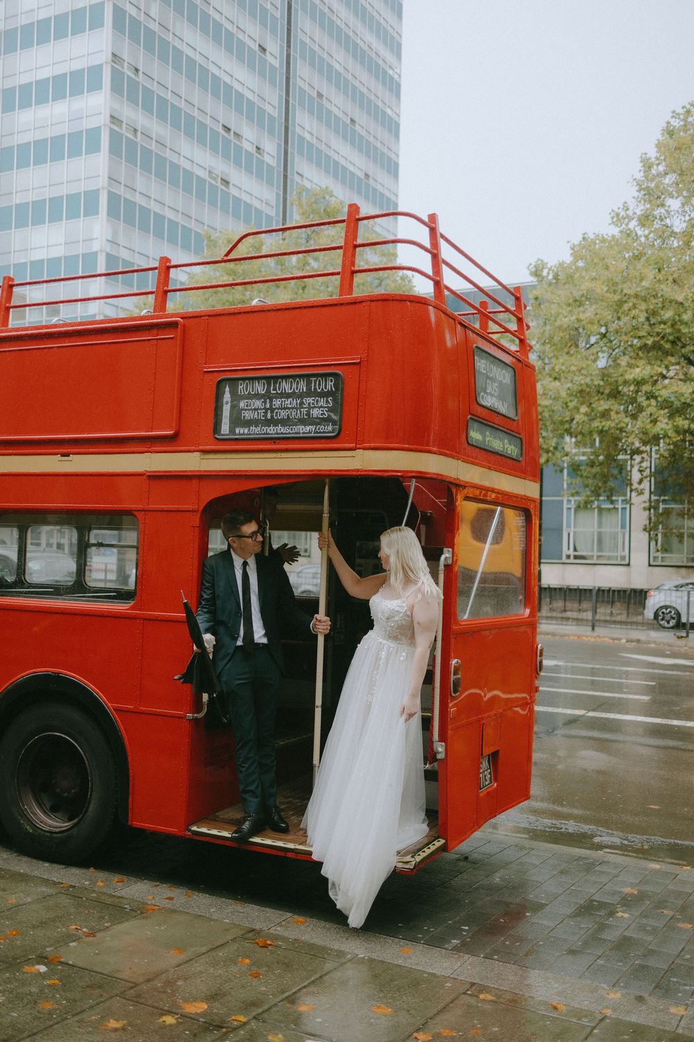Bride in white wedding dress boarding a classic red London double-decker bus with groom.