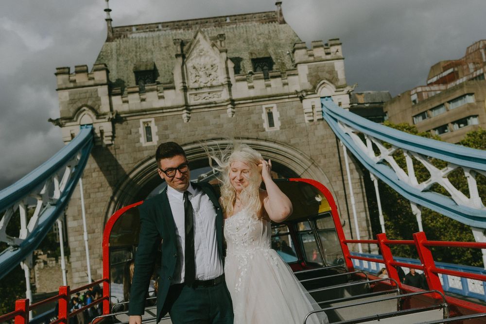 Wedding couple celebrates joyfully on Tower Bridge in London with cloudy skies overhead.
