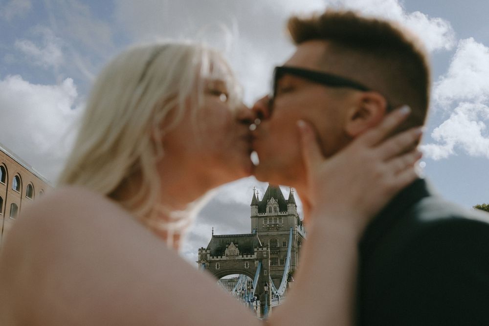 Romantic couple shares an intimate kiss in London with Tower Bridge visible in the background against cloudy skies.