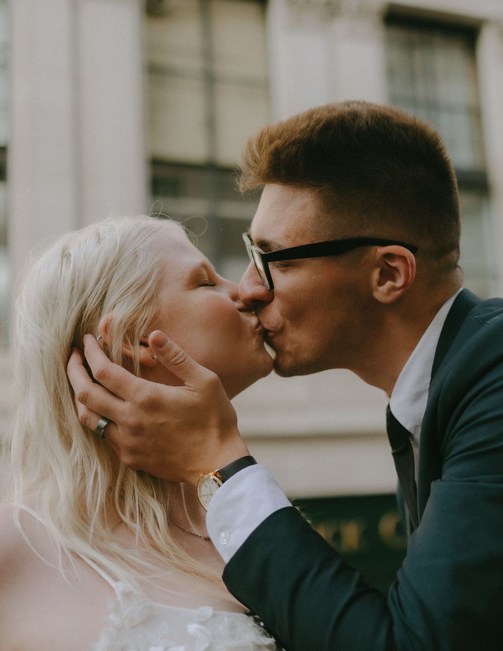 A tender wedding kiss between bride in white dress and groom in glasses during an intimate outdoor ceremony.